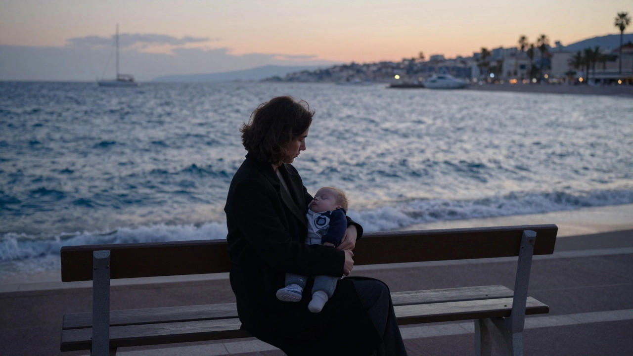 A woman sits on a Cannes beach bench with a sleeping baby in her lap as the sun sets over the ocean.